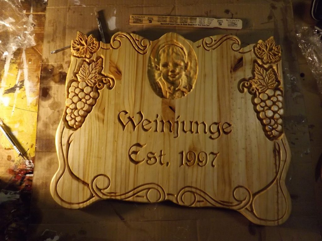 A wooden plaque engraved with grapevine designs, a child's portrait, and the text "Weinjunge Est. 1997".