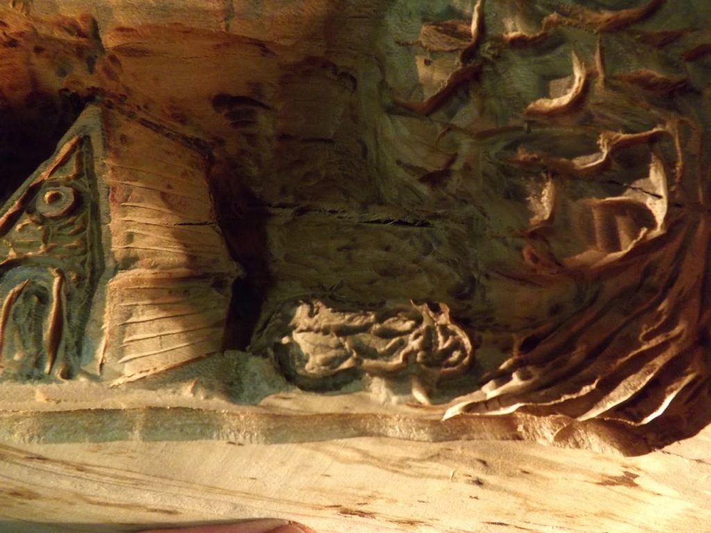 Wood carving depicting a small house, abstract shapes, and textured patterns in a rustic style.