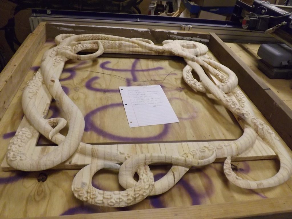 A wooden picture frame carved with an octopus tentacle design sits on a workbench with a handwritten note in the center.