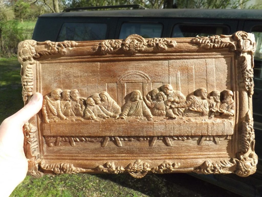 A hand holds a carved wooden plaque depicting the scene of The Last Supper in front of a vehicle.