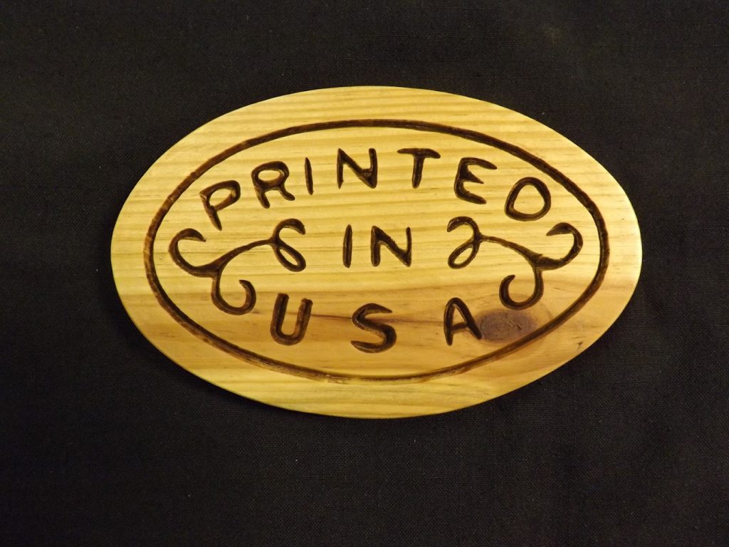 Oval wooden plaque with the words "Printed in USA" engraved, placed on a plain black background.
