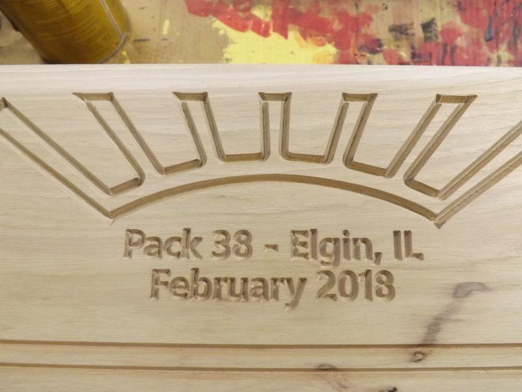 Wood carving reads "Pack 38 - Elgin, IL February 2018" with a sunburst design above the text.
