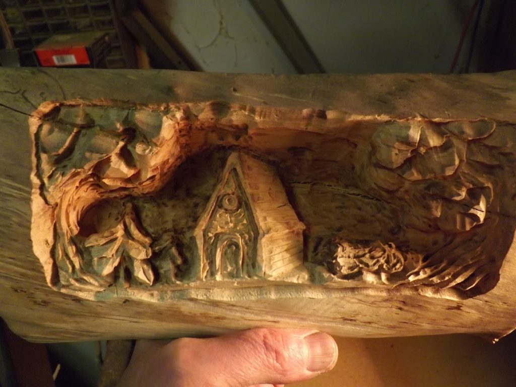 A hand holds a carved wooden scene depicting a small house, trees, and bushes in a forest setting.