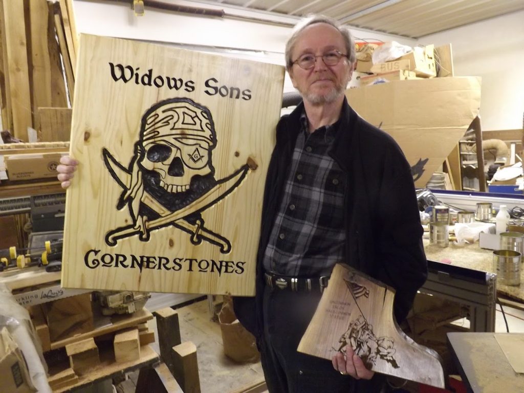 An older man holds a wooden sign reading "Widows Sons Cornerstones" with a skull and crossbones design in a workshop.
