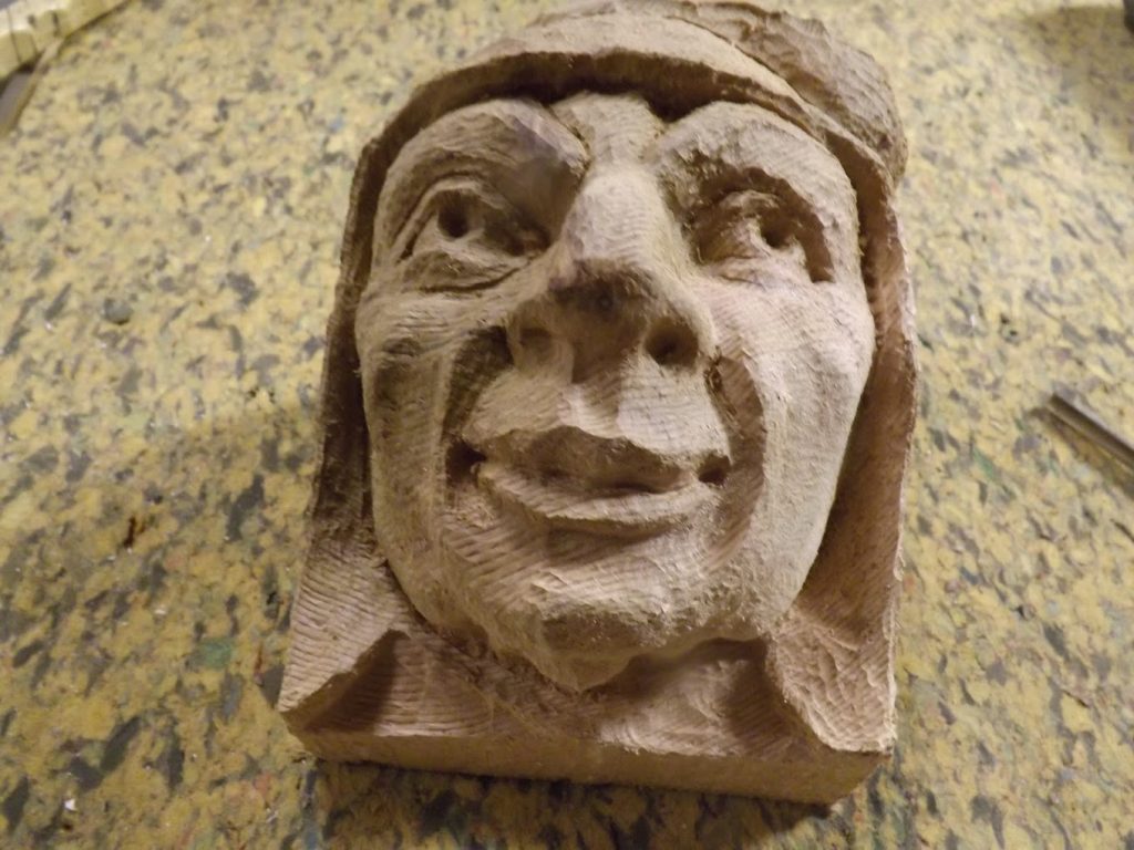 A roughly carved wooden face with exaggerated features, lying on a speckled surface.